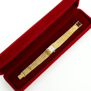 May include: A gold-toned wristwatch with a textured band and a square face, presented in a red velvet box. The watch features a rectangular dial with a light-colored face. The band has a textured, woven appearance.