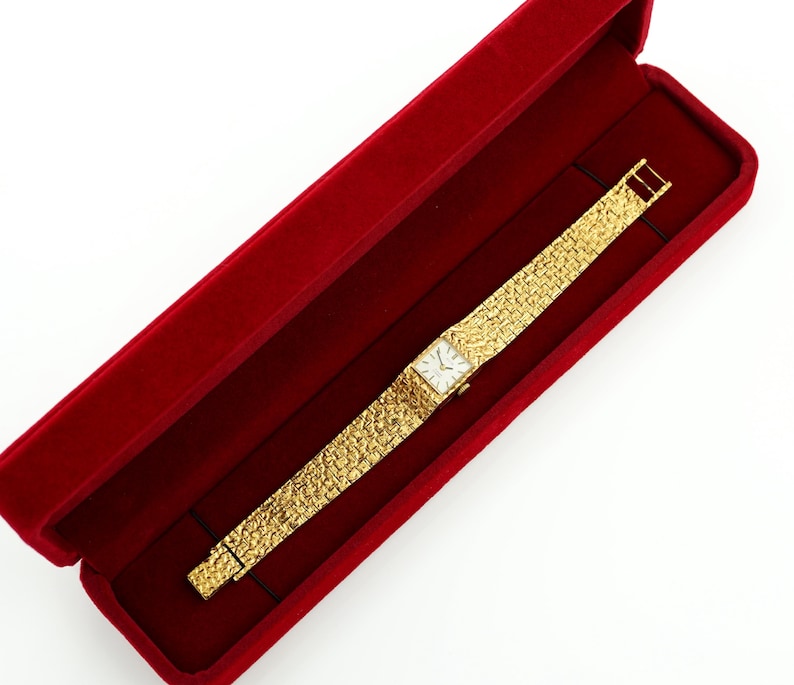 May include: A gold-toned wristwatch with a textured band and a square face, presented in a red velvet box. The watch features a rectangular dial with a light-colored face. The band has a textured, woven appearance.