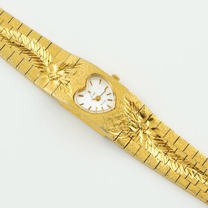 May include: A gold-tone quartz watch with a heart-shaped white dial featuring black hour markers and hands. The integrated metal bracelet has a textured finish with engraved leaf-like patterns on both sides of the watch case.