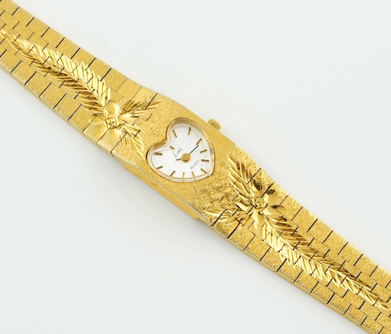 May include: A gold-tone quartz watch with a heart-shaped white dial featuring black hour markers and hands. The integrated metal bracelet has a textured finish with engraved leaf-like patterns on both sides of the watch case.