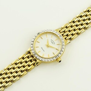Puede incluir: Un reloj Rotary dorado con una esfera blanca y un bisel engastado con diamantes. El reloj tiene una pulsera de metal dorado.