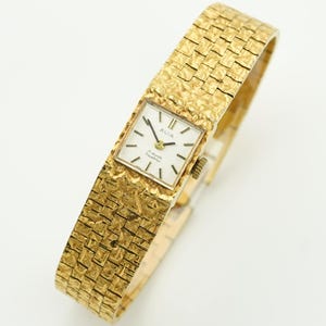 May include: A gold-toned Avia wristwatch with a textured band and square face. The watch face is white with black hands and markers. The band has a woven appearance. The watch is a vintage style.