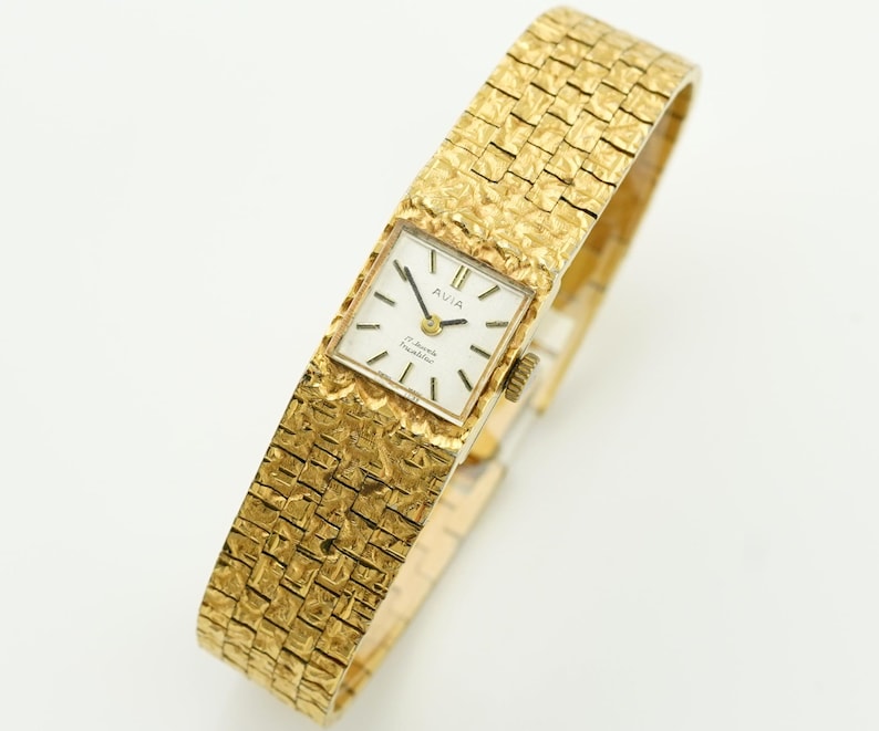 May include: A gold-toned Avia wristwatch with a textured band and square face. The watch face is white with black hands and markers. The band has a woven appearance. The watch is a vintage style.