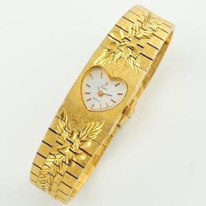 May include: A gold-tone quartz wristwatch featuring a heart-shaped white dial with the brand name Limit printed on the face. The watch has a textured gold-tone metal link bracelet adorned with embossed leaf patterns above and below the dial.