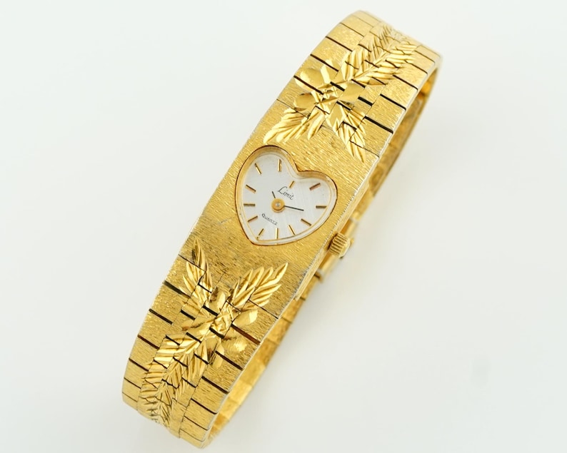 May include: A gold-tone quartz wristwatch featuring a heart-shaped white dial with the brand name Limit printed on the face. The watch has a textured gold-tone metal link bracelet adorned with embossed leaf patterns above and below the dial.