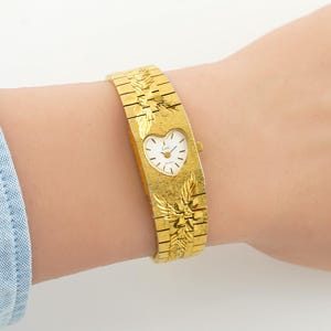 May include: A gold-tone wristwatch featuring a wide, textured metal band with engraved leaf patterns. The watch face is heart-shaped with a white dial, gold-tone hands, and the brand name Lini printed in black. A small crown is on the right side.