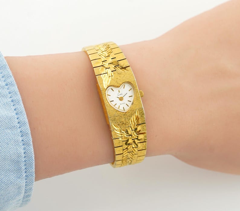 May include: A gold-tone wristwatch featuring a wide, textured metal band with engraved leaf patterns. The watch face is heart-shaped with a white dial, gold-tone hands, and the brand name Lini printed in black. A small crown is on the right side.