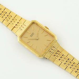 May include: A gold-toned Seiko Quartz watch with a rectangular face and a matching textured band. The watch face is a light gold color with simple hour markers and hands. The band features a repeating geometric pattern.