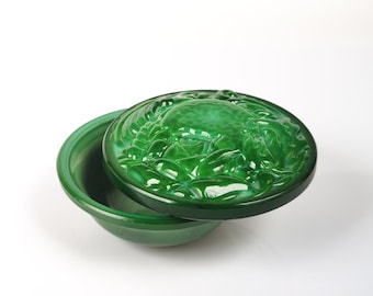 Puede incluir: Una caja de vidrio verde para baratijas con una tapa que presenta un diseño en relieve de un motivo floral estilizado. La caja está hecha de vidrio verde translúcido con un acabado brillante.