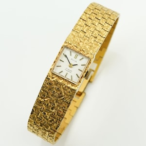 May include: A gold-toned Avia wristwatch with a textured band and square face. The watch face is white with black hands and markers. The band has a woven pattern. The watch is displayed on a white surface.