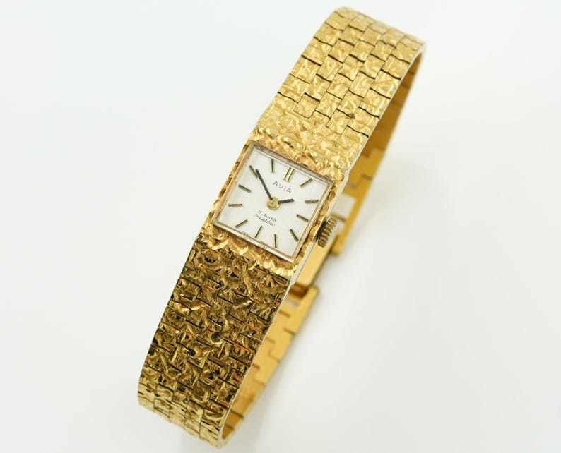 May include: A gold-toned Avia wristwatch with a textured band and square face. The watch face is white with black hands and markers. The band has a woven pattern. The watch is displayed on a white surface.