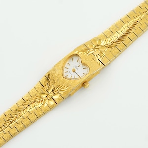 May include: A gold-tone wristwatch featuring a heart-shaped white dial with gold-tone hands and markers. The watch has a textured gold-tone metal link bracelet with an engraved leaf-like pattern extending from the case onto the band.