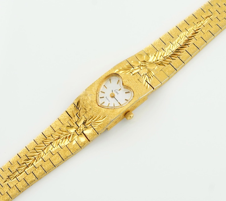 May include: A gold-tone wristwatch featuring a heart-shaped white dial with gold-tone hands and markers. The watch has a textured gold-tone metal link bracelet with an engraved leaf-like pattern extending from the case onto the band.