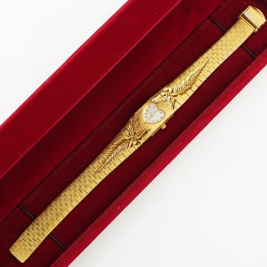 May include: A gold-tone wristwatch with a heart-shaped white dial and a textured link bracelet. The watch features decorative leaf-like engravings on the band surrounding the dial. It is presented inside a rectangular red velvet-lined display case.