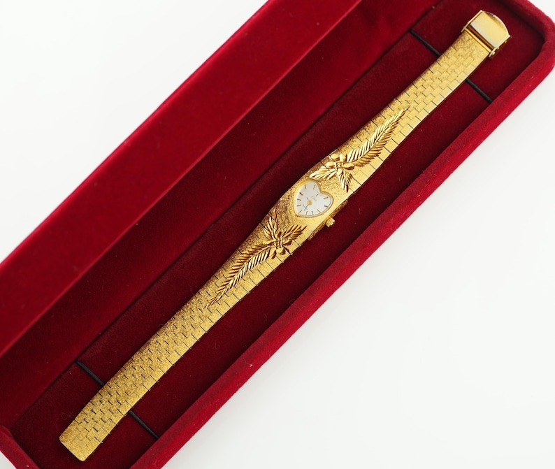 May include: A gold-tone wristwatch with a heart-shaped white dial and a textured link bracelet. The watch features decorative leaf-like engravings on the band surrounding the dial. It is presented inside a rectangular red velvet-lined display case.