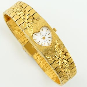 May include: A gold-tone quartz wristwatch featuring a heart-shaped white dial with the text 'Limi' printed on the face. The watch has a wide, textured metal band with an engraved leaf pattern on both sides of the central heart-shaped watch case.