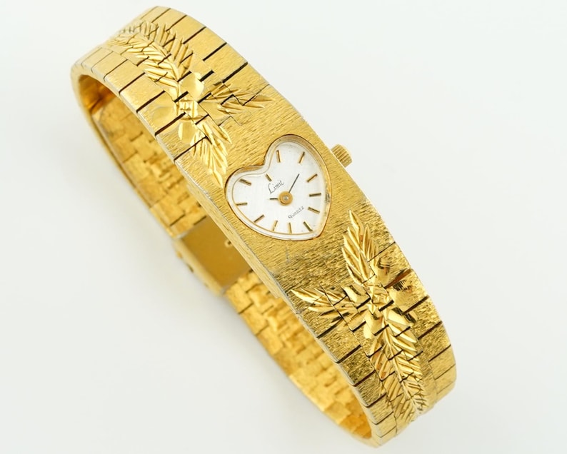 May include: A gold-tone quartz wristwatch featuring a heart-shaped white dial with the text 'Limi' printed on the face. The watch has a wide, textured metal band with an engraved leaf pattern on both sides of the central heart-shaped watch case.