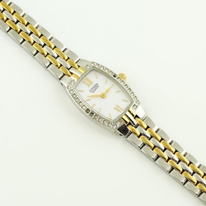 May include: A Citizen Quartz wristwatch with a white face, gold hands, and a silver and gold band. The watch face is surrounded by small, clear crystals. The band is a combination of silver and gold links.