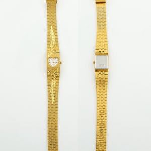 May include: A gold-tone wristwatch featuring a heart-shaped dial with a white face and gold hands. The band is a textured gold-tone metal link strap with embossed feather-like designs above and below the dial. The back of the watch case is silver.