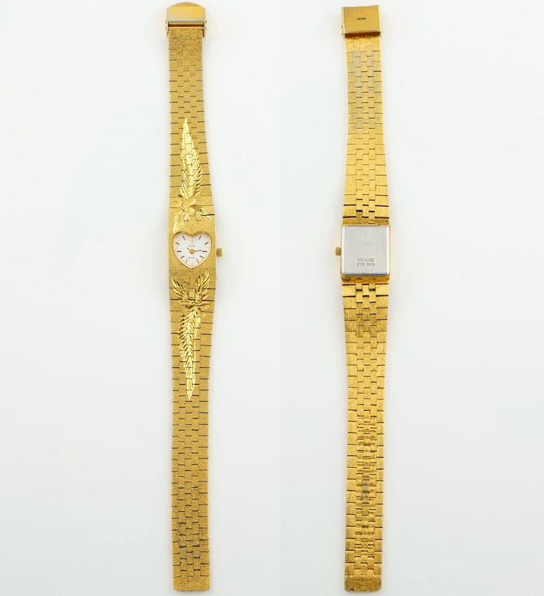 May include: A gold-tone wristwatch featuring a heart-shaped dial with a white face and gold hands. The band is a textured gold-tone metal link strap with embossed feather-like designs above and below the dial. The back of the watch case is silver.