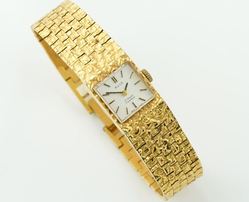 May include: A gold-toned Avia wristwatch with a textured band and square face. The watch face is white with black hands and markers. The band has a woven pattern. The watch is displayed on a white background.