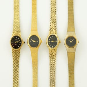 Può includere: Quattro orologi da polso dorati con quadranti neri sono esposti. Gli orologi hanno diversi stili di cinturino, tra cui modelli a maglia e a maglie. I quadranti hanno lancette e indicatori dorati.