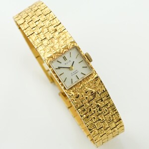 May include: A vintage gold-toned Avia wristwatch with a textured band. The watch face is square with black hands and markers. The band has a woven pattern. The watch is a classic accessory.