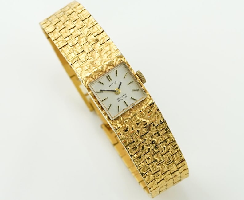 May include: A vintage gold-toned Avia wristwatch with a textured band. The watch face is square with black hands and markers. The band has a woven pattern. The watch is a classic accessory.