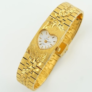 May include: A gold-tone watch with a textured finish and a heart-shaped white dial. The watch features a wide, integrated link bracelet with engraved leaf-like patterns above and below the dial. The dial displays the text "Lenz" and "Quartz".