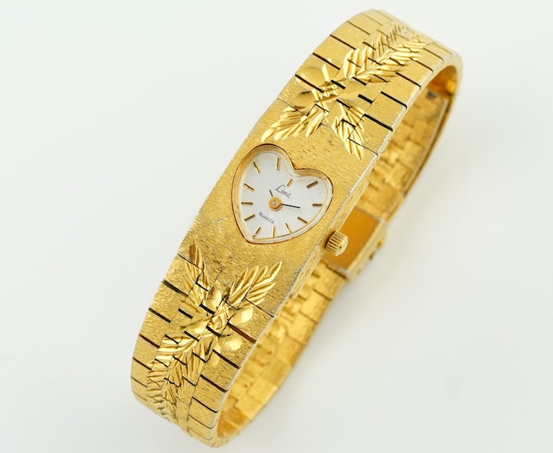 May include: A gold-tone watch with a textured finish and a heart-shaped white dial. The watch features a wide, integrated link bracelet with engraved leaf-like patterns above and below the dial. The dial displays the text "Lenz" and "Quartz".