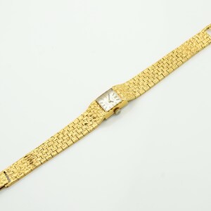 May include: A gold-toned wristwatch with a rectangular face and a textured band. The watch face is white with black hands and markers. The band is made of small, textured gold links. The watch is displayed on a white surface.