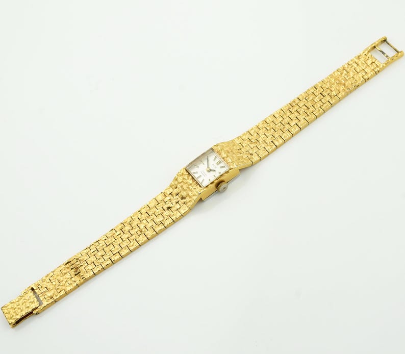 May include: A gold-toned wristwatch with a rectangular face and a textured band. The watch face is white with black hands and markers. The band is made of small, textured gold links. The watch is displayed on a white surface.