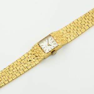 May include: A gold-toned wristwatch with a textured band and square face. The watch face is white with black hands and markers. The band has a woven pattern. The watch is set against a white background.