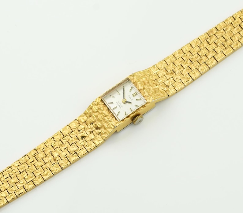 May include: A gold-toned wristwatch with a textured band and square face. The watch face is white with black hands and markers. The band has a woven pattern. The watch is set against a white background.