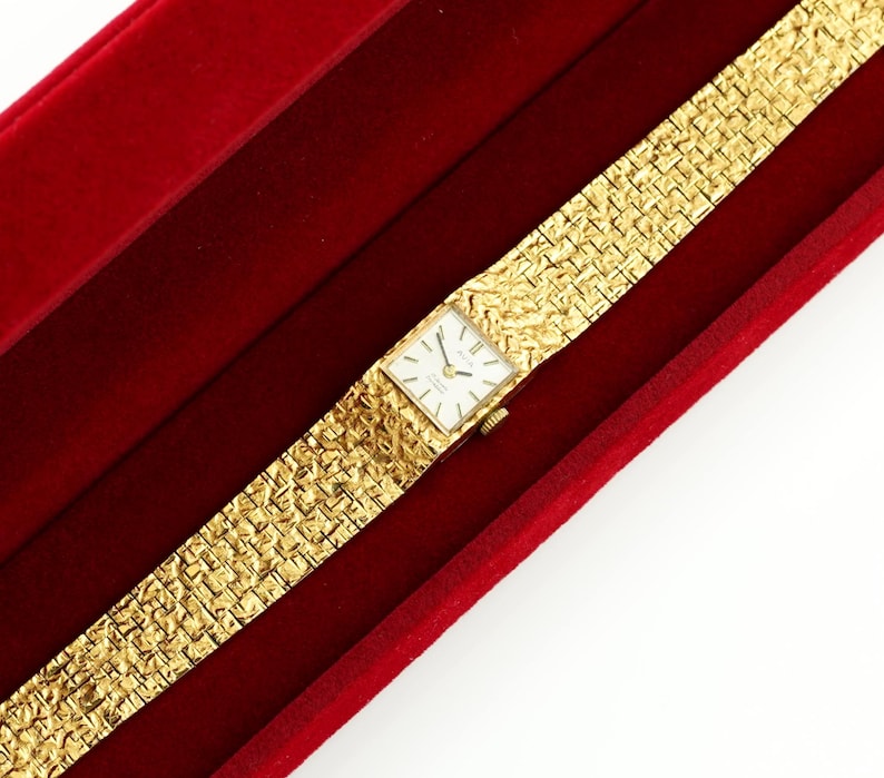 May include: A gold-toned wristwatch with a rectangular face and a textured band, resting in a red velvet box. The watch face is white with black hands and markers. The band has a woven pattern.