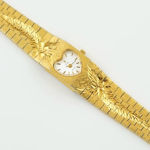 May include: A gold-tone wristwatch featuring a heart-shaped white dial with black hour markers and hands. The watch has a textured gold-tone metal link bracelet with integrated leaf-like engravings on the band. The dial displays the text Limit.