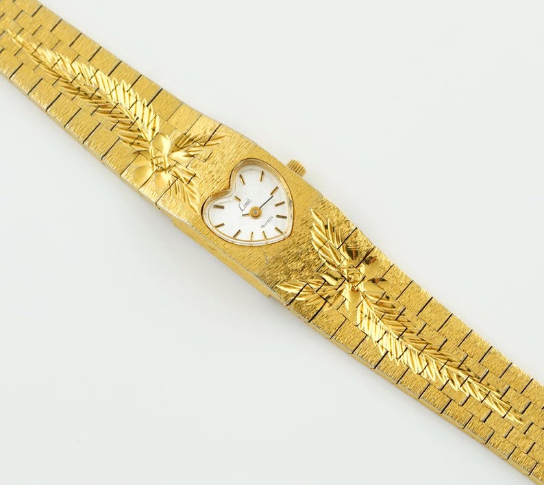 May include: A gold-tone wristwatch featuring a heart-shaped white dial with black hour markers and hands. The watch has a textured gold-tone metal link bracelet with integrated leaf-like engravings on the band. The dial displays the text Limit.