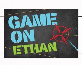 May include: A printable sign with a black chalkboard background. The sign says "Game On Ethan" in blue and green letters with a red target symbol. The sign is perfect for a birthday party or other celebration.