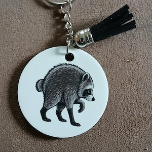May include: A circular white keychain featuring a detailed black and gray illustration of a raccoon walking. The keychain includes a silver metal split ring, a short silver chain, and a small black faux suede tassel attached to the top.