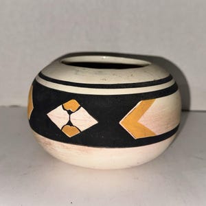 Op de afbeelding: Een kleine, ronde, crèmekleurige keramische pot met een brede horizontale band van matte zwarte verf. De zwarte band is versierd met geometrische vormen in mosterdgeel en lichtroze, omlijst door dunne zwarte lijnen rond de rand en basis.