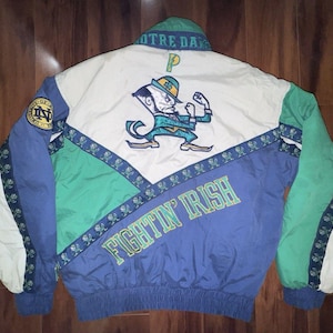 Puede incluir: Una chaqueta Notre Dame Fighting Irish de bloques de color blanco, azul y verde. La espalda presenta una mascota de duende bordada, el texto "FIGHTIN' IRISH" en amarillo, un cuello "NOTRE DAME" y una banda con tréboles.
