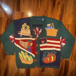 May include: Green cardigan with embroidered images of a sunflower cart, a basket with a teddy bear, a basket of fruit, an American flag, and pumpkins. The sweater has red buttons and a V-neck.