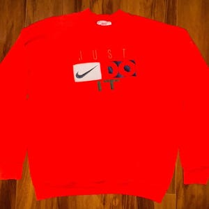 May include: Bright red sweatshirt with the Nike logo and the words "JUST DO IT" in a white, blue, and green design. The sweatshirt has long sleeves and a crew neck. The Nike logo is a white rectangle with a black checkmark.