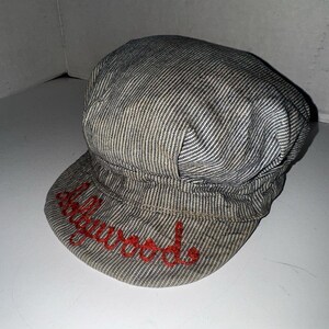 May include: A grey and white striped newsboy cap with a short peak. The word "Hollywood" is embroidered in red script across the front. The cap has a rounded crown and a classic design, suitable for various outfits.