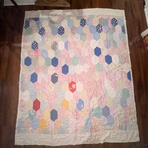 Op de afbeelding: Een rechthoekige quilt met een patchwork van zeshoekige stoffen stukjes in diverse pastel- en primaire kleuren, waaronder roze, blauw, geel en rood. De quilt is aan alle vier de zijden afgezet met een effen, gebroken witte stoffen rand.