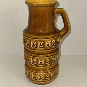 May include: A brown ceramic pitcher with a handle. The pitcher has a cylindrical body with a series of horizontal bands decorated with square and circle patterns. The top and handle are a lighter shade of brown.