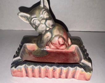 May include: A ceramic ashtray featuring a sculpted French bulldog figure attached to the back. The piece has a rectangular base with a fluted edge, finished in a mottled glaze of dusty pink, muted green, and charcoal gray tones throughout.