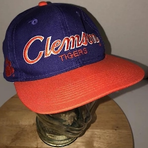 Puede incluir: Gorra de béisbol azul y naranja con la palabra "Clemson" bordada en escritura naranja en la parte delantera. La palabra "Tigers" está bordada debajo. La gorra tiene una visera naranja curvada y un pequeño botón naranja en la parte superior.