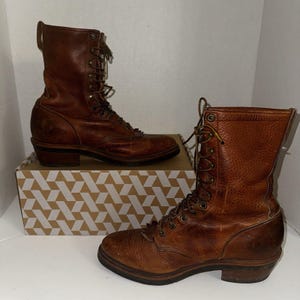 Może przedstawiać: Para brązowych skórzanych butów sznurowanych. Buty mają teksturowany wygląd, wysokość do połowy łydki i niski obcas. Sznurowadła są brązowe, a buty są prezentowane na pudełku ze wzorem.