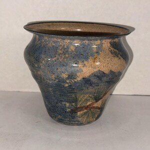 以下が含まれることがあります： 広いフレア状の縁と先細りの底を持つ陶器製のプランター。外側はベージュと青の斑点のある釉薬で仕上げられており、濃い青色の山のシルエットと緑の針葉を持つ茶色の松の枝が描かれた風景画が施されています。